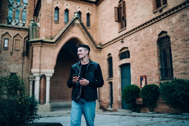 Contenu souriant hipster homme adulte en veste de jeans debout avec smartphone et casque