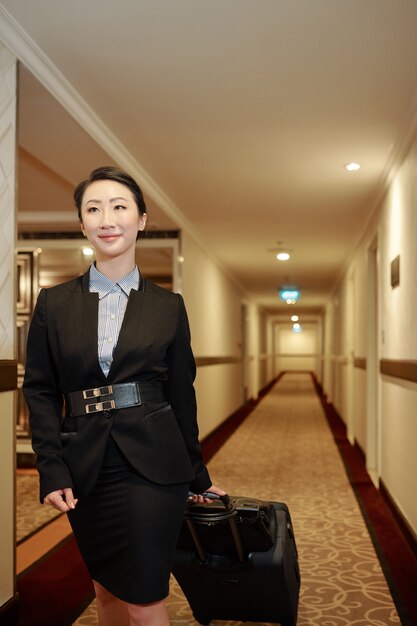 Photo contetn jolie jeune femme asiatique en tenue de soirée étant en voyage d'affaires marchant avec des bagages le long du couloir de l'hôtel