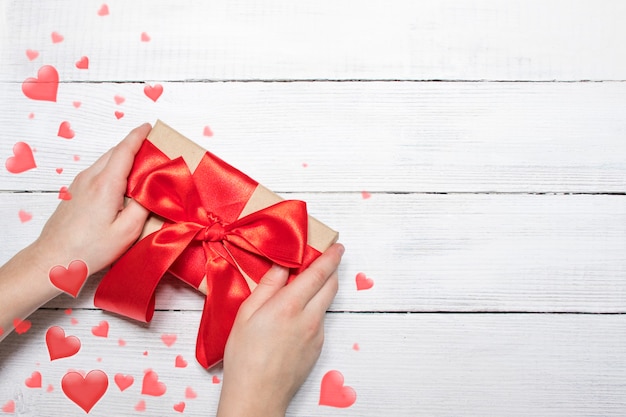 Contexte de la Saint-Valentin. Mains tiennent une boîte avec un cadeau et un ruban rouge sur un fond en bois blanc