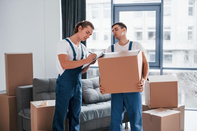 Contient un bloc-notes avec un document Deux jeunes déménageurs en uniforme bleu travaillant à l'intérieur dans la chambre