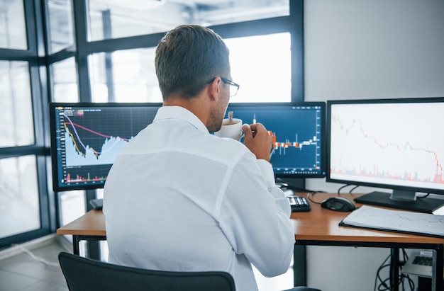 Contient une tasse de boisson. Analyser les informations. Un jeune homme d'affaires en vêtements formels est en poste avec plusieurs écrans. Conception de l'échange et de la monnaie.