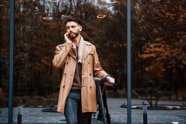 Contre le bâtiment et avec un scooter électrique Jeune modèle masculin dans des vêtements à la mode est à l'extérieur dans la ville pendant la journée