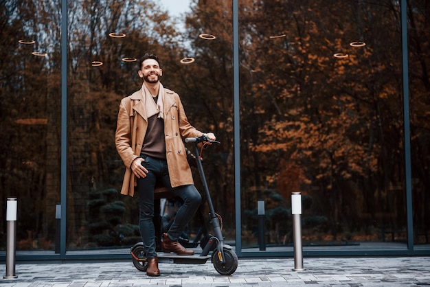 Contre le bâtiment et avec un scooter électrique Jeune modèle masculin dans des vêtements à la mode est à l'extérieur dans la ville pendant la journée