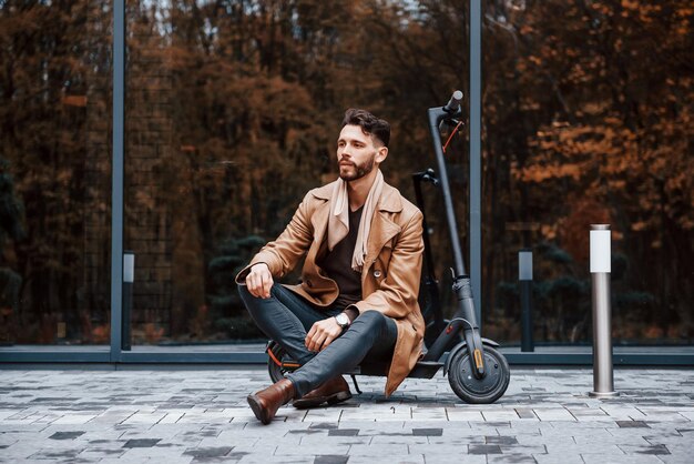 Contre le bâtiment et avec un scooter électrique Jeune modèle masculin dans des vêtements à la mode est à l'extérieur dans la ville pendant la journée