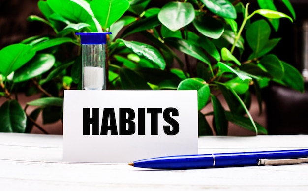 Contre la surface des feuilles vertes de la plante, il y a un stylo sur la table, un sablier et une carte avec l'inscription HABITS