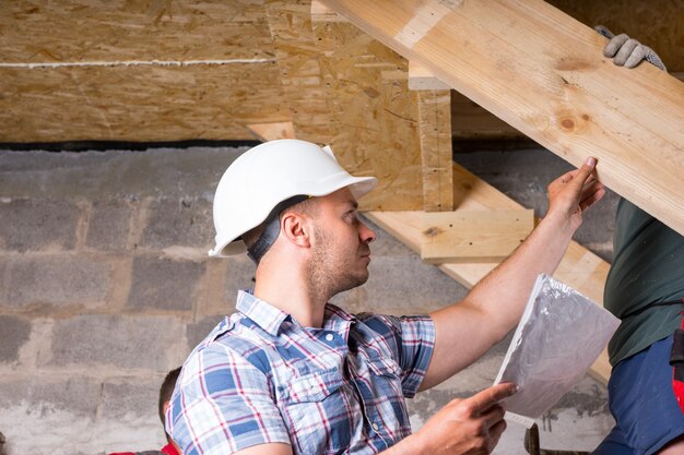 Contremaître de chantier portant un casque blanc et tenant des plans de construction inspectant le travail d'équipe sur un escalier en bois dans un sous-sol inachevé d'une nouvelle maison