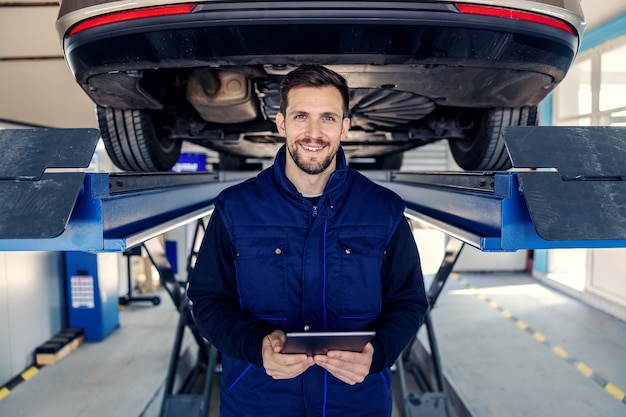 Contrôle technique des voitures et des tablettes. Un homme en uniforme tient une tablette dans ses mains dans le canal de l'atelier devant une voiture sur un ascenseur hydraulique Portrait d'un homme au travail dans un atelier Service de voiture