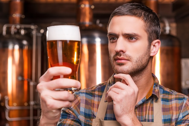 Contrôler la qualité de la bière. Jeune brasseur réfléchi en tablier tenant un verre avec de la bière et le regardant tout en se tenant devant des récipients en métal
