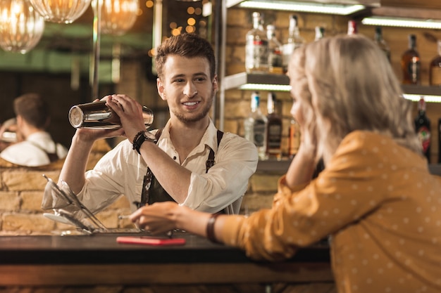 Conversation polie. Agréable jeune barman faisant un cocktail et souriant à son client pendant qu'elle lui parle