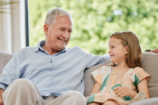 Photo conversation sourire et grand-père avec l'enfant sur un canapé l'attachement de relaxation et de parler à la maison famille heureuse et homme âgé en train de parler à une jeune fille enfant sur un divan dans le salon à la maison moderne