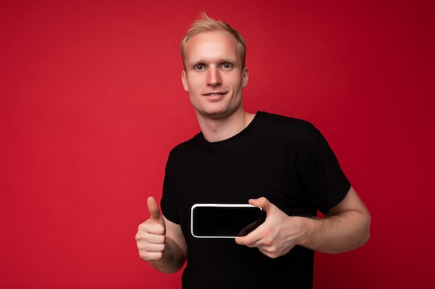 Cool beau jeune homme blond sérieux portant un tshirt noir debout isolé sur fond rouge avec