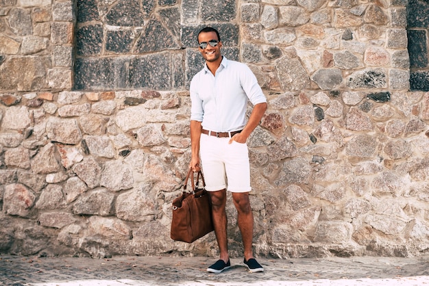 Cool et beau. Toute la longueur d'un jeune homme africain joyeux dans des vêtements décontractés et élégants, portant un sac et regardant la caméra en se tenant debout contre le mur de pierres à l'extérieur