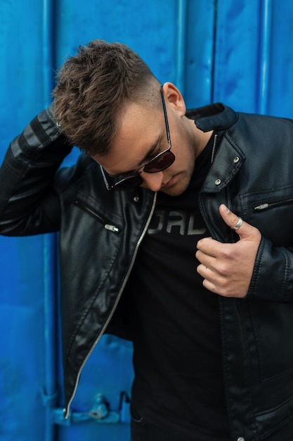 Cool branché jeune hipster homme avec une coiffure en tenue noire avec une veste en cuir noire et des lunettes de soleil de mode debout à côté d'un mur en métal vintage bleu