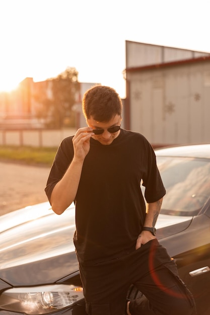 Cool fashion hipster homme avec des lunettes de soleil en t-shirt noir et jeans se tient près d'une voiture au coucher du soleil