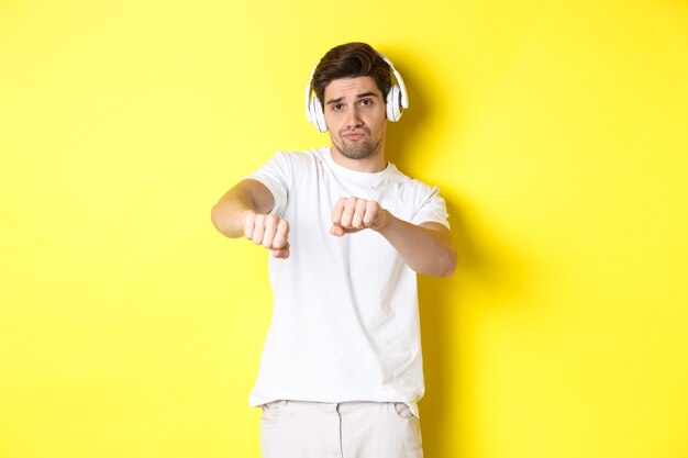 Cool guy écoutant de la musique dans les écouteurs et dansant, debout dans des vêtements blancs contre le mur jaune du studio