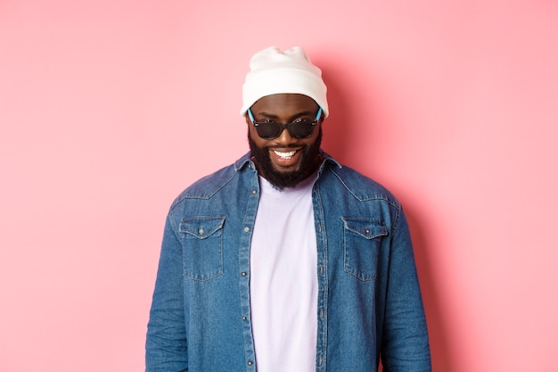 Cool hipster afro-américain en bonnet et lunettes de soleil, souriant heureux et regardant la caméra, debout confiant sur fond rose