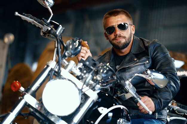 Cool homme barbu motard à lunettes de soleil assis sur sa moto et fumer le cigare