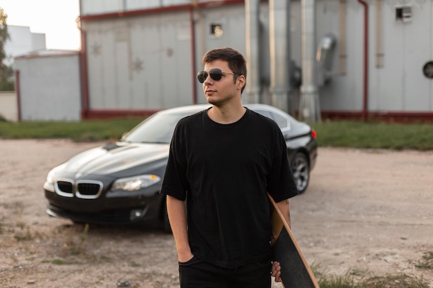 Cool homme hipster élégant avec des lunettes de soleil de mode dans un t-shirt noir et un jean avec un longboard se promène sur le fond d'une voiture Activités de plein air et week-ends