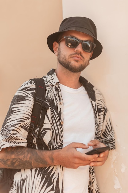 Cool jeune bel homme hipster avec des lunettes de soleil de mode et un chapeau en chemise d'été avec sac à dos avec smartphone marche dans la rue Style masculin d'été urbain