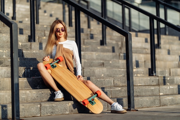 Cool jeune femme avec un longboard assis sur les escaliers à lunettes de soleil. Style décontracté. Photo de haute qualité