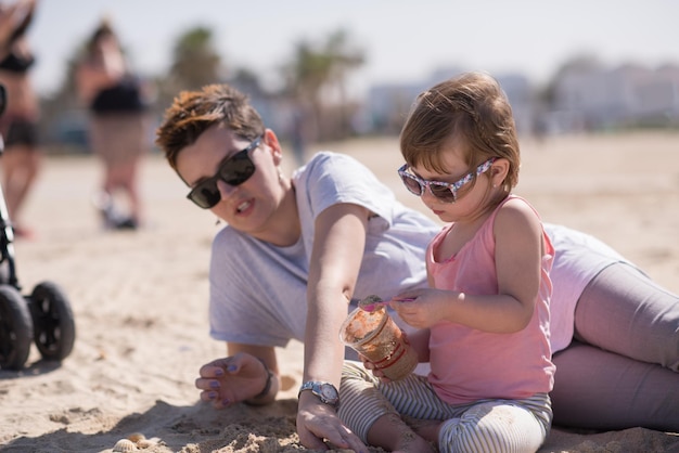 cool jeune maman et sa petite fille jouant avec un seau plein de sable portant des lunettes de soleil et se relaxant sur la plage par une journée ensoleillée