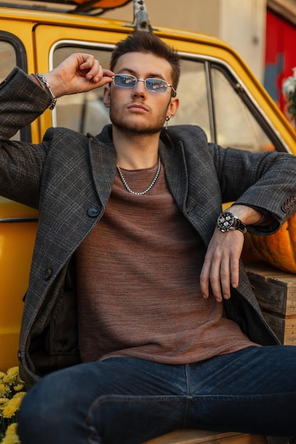 Cool à la mode jeune homme hipster avec un jean manteau à la mode et un sweat-shirt porte des lunettes de soleil et se repose près d'une voiture rétro jaune