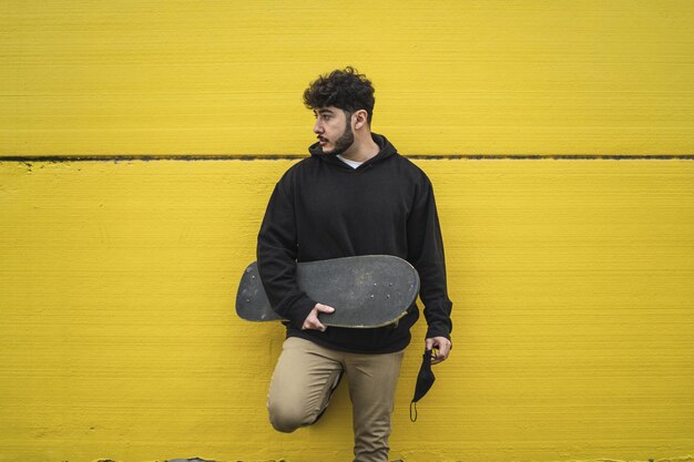 Cool skateboarder masculin européen s'appuyant sur un mur jaune vif