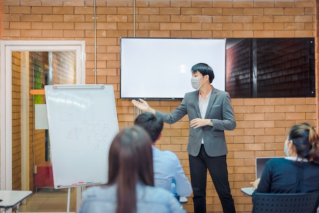 Coopération commerciale : un jeune entraîneur ou conférencier asiatique fait une présentation sur un tableau à feuilles mobiles à divers hommes d'affaires lors d'une réunion au bureau. Un tuteur ou un formateur masculin présente le projet à divers collègues.