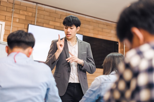 Coopération commerciale : un jeune entraîneur ou conférencier asiatique fait une présentation sur un tableau à feuilles mobiles à divers hommes d'affaires lors d'une réunion au bureau. Un tuteur ou un formateur masculin présente le projet à divers collègues.