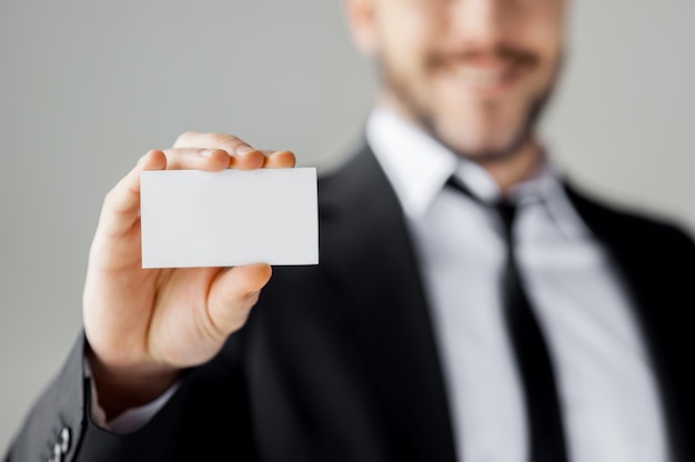 Copiez l'espace sur sa carte de visite. Gai jeune homme en tenue de soirée montrant sa carte de visite et souriant en se tenant debout sur fond gris