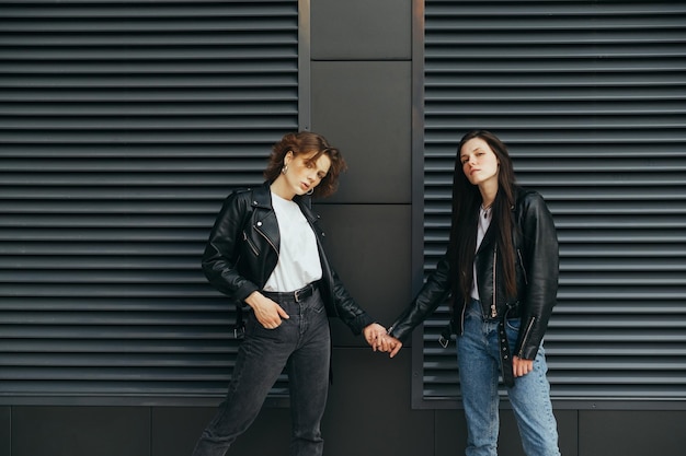 Des copines élégantes se tiennent la main contre un mur sombre et posent pour la caméra