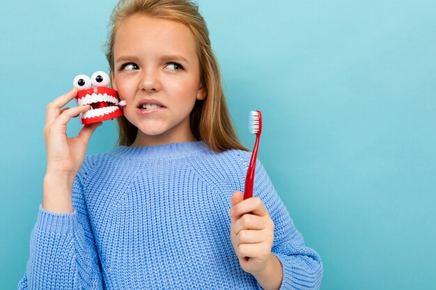 Copyspace photo de charmante écolière pensive avec une brosse à dents et une maquette dentaire sur fond de mur bleu
