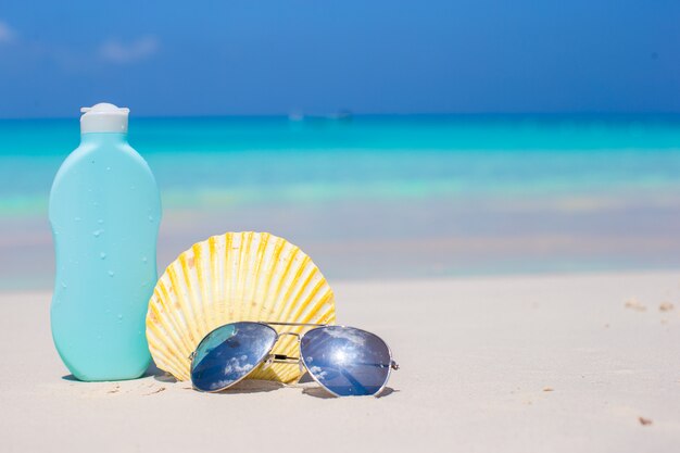 Coquillage, lunettes de soleil et crème solaire sur le sable blanc