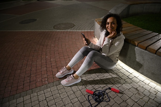 Une corde à sauter allongée sur le sol à côté d'une belle femme afro-américaine s'appuyant sur un banc tenant un téléphone portable et un joli sourire en regardant la caméra