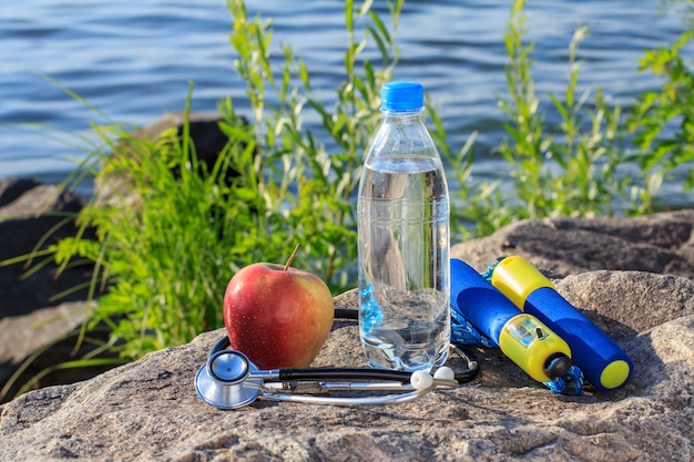 Corde à sauter, pomme, stéthoscope et bouteille d'eau sur un rocher avec fond de remblai de rivière. Été, mode de vie actif