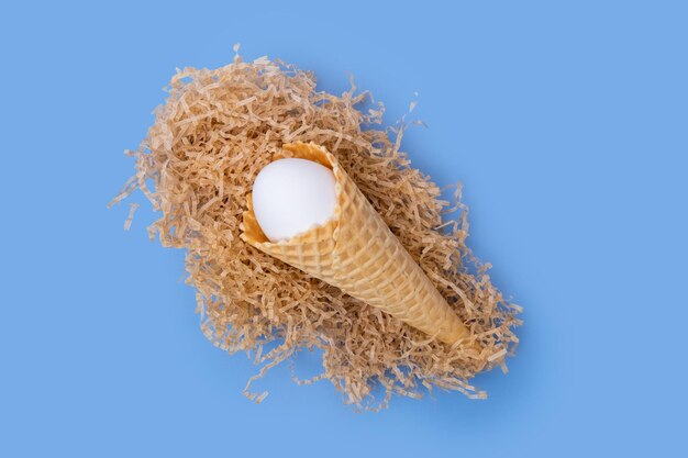 Cornet de crème glacée gaufré avec un œuf de Pâques blanc caché dans un nid de papier sur fond bleu