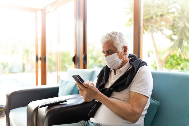 Coronavirus Covid-19 Homme âgé âgé à La Maison S'asseoir Sur Le Canapé Dans Un Masque Médical. Surveiller Son Téléphone Pendant La Pandémie.restez à La Maison