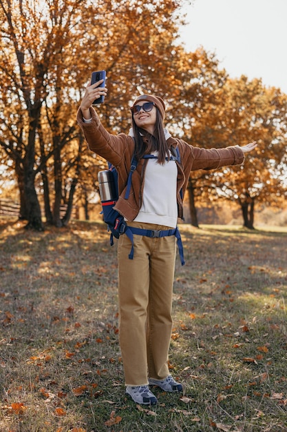 Corps complet d'une femme voyageuse heureuse avec sac à dos prenant selfie sur smartphone dans les bois d'automne