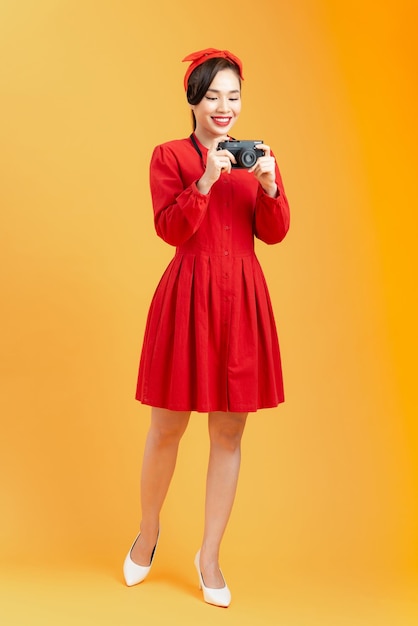 Corps entier d'une belle jeune femme asiatique vêtue d'une robe rouge en studio sur fond orange avec un appareil photo dans les mains