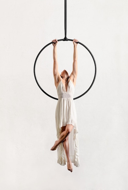 Corps entier de femme acrobatique pieds nus active en robe suspendue à un cerceau aérien tout en effectuant des exercices sur fond blanc en studio
