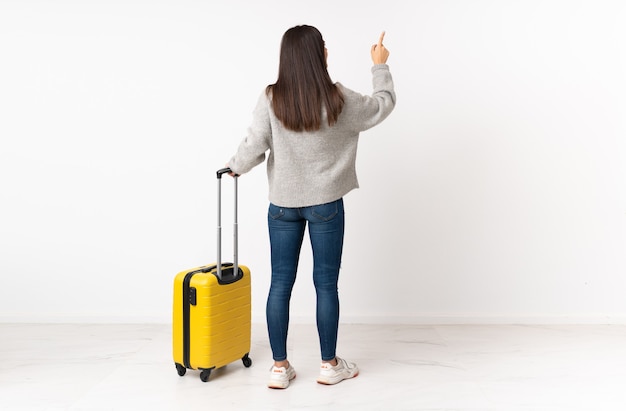 Un corps entier d'une femme voyageur avec une valise sur un mur blanc isolé pointant vers l'arrière avec l'index