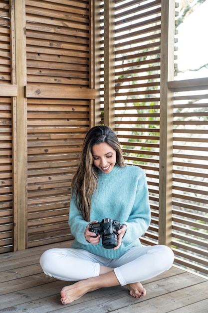 Corps entier de joyeuse jeune femme aux pieds nus dans des vêtements décontractés avec appareil photo en mains assis sur une terrasse en bois