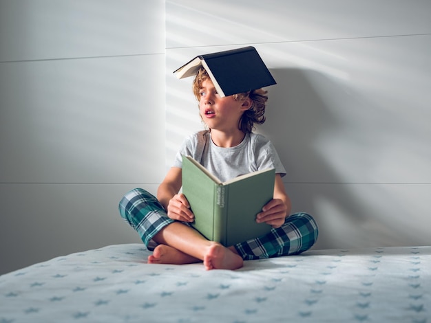 Corps entier d'un mignon petit garçon fatigué en pyjama assis sur le lit avec un manuel ouvert et une tête couverte de livre
