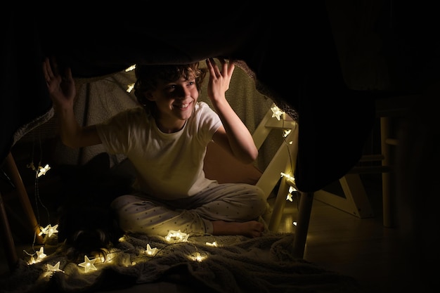 Un corps plein d'un garçon joyeux détournant le regard alors qu'il est assis sous une construction de couverture avec une guirlande brillante dans une pièce sombre à la maison