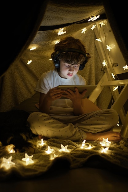 Corps plein de garçon sérieux dans des écouteurs regardant une vidéo sur une tablette moderne tout en étant assis avec les jambes croisées près d'une guirlande brillante dans une pièce sombre