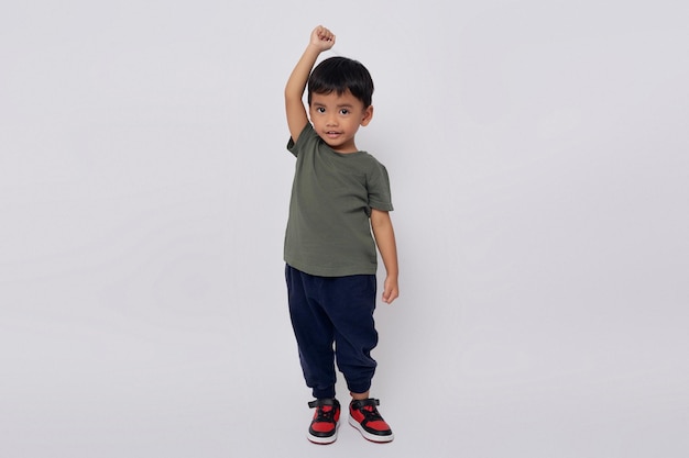 Photo corps plein petit petit sourire heureux garçon asiatique de 23 ans portant un t-shirt vert debout confiant tout en faisant le geste du vainqueur isolé sur fond blanc