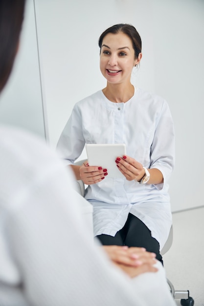 Cosméticienne caucasienne attentive et attrayante avec une tablette informatique assise devant son patient