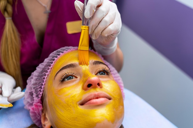 Cosmétologue appliquant un masque d'or sur le visage de la femme à l'aide d'une brosse en cosmétologie Portrait d'une jeune fille sur la procédure de nettoyage de la peau par une esthéticienne dans une clinique de beauté Photo du haut