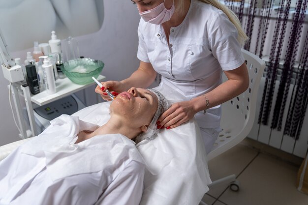 Le cosmétologue applique le gel sur le visage du patient, en le répartissant uniformément sur la peau avec une brosse spéciale