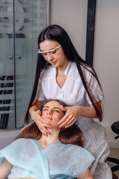 Une cosmétologue féminine lisse les rides de son visage avec ses mains à une fille allongée sur un canapé dans un salon de beauté.